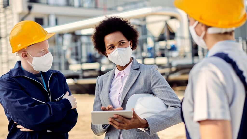 Black businesswoman and two construction workers wearing protective face masks while talking about ongoing project outdoors.